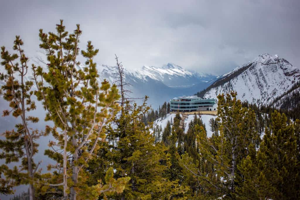 a building sits atop a mountain with spruce trees in the foreground