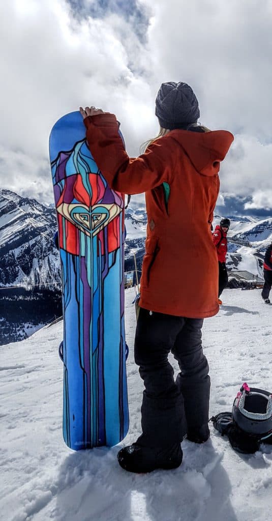 girl stands with her snowboard on top of a mountain