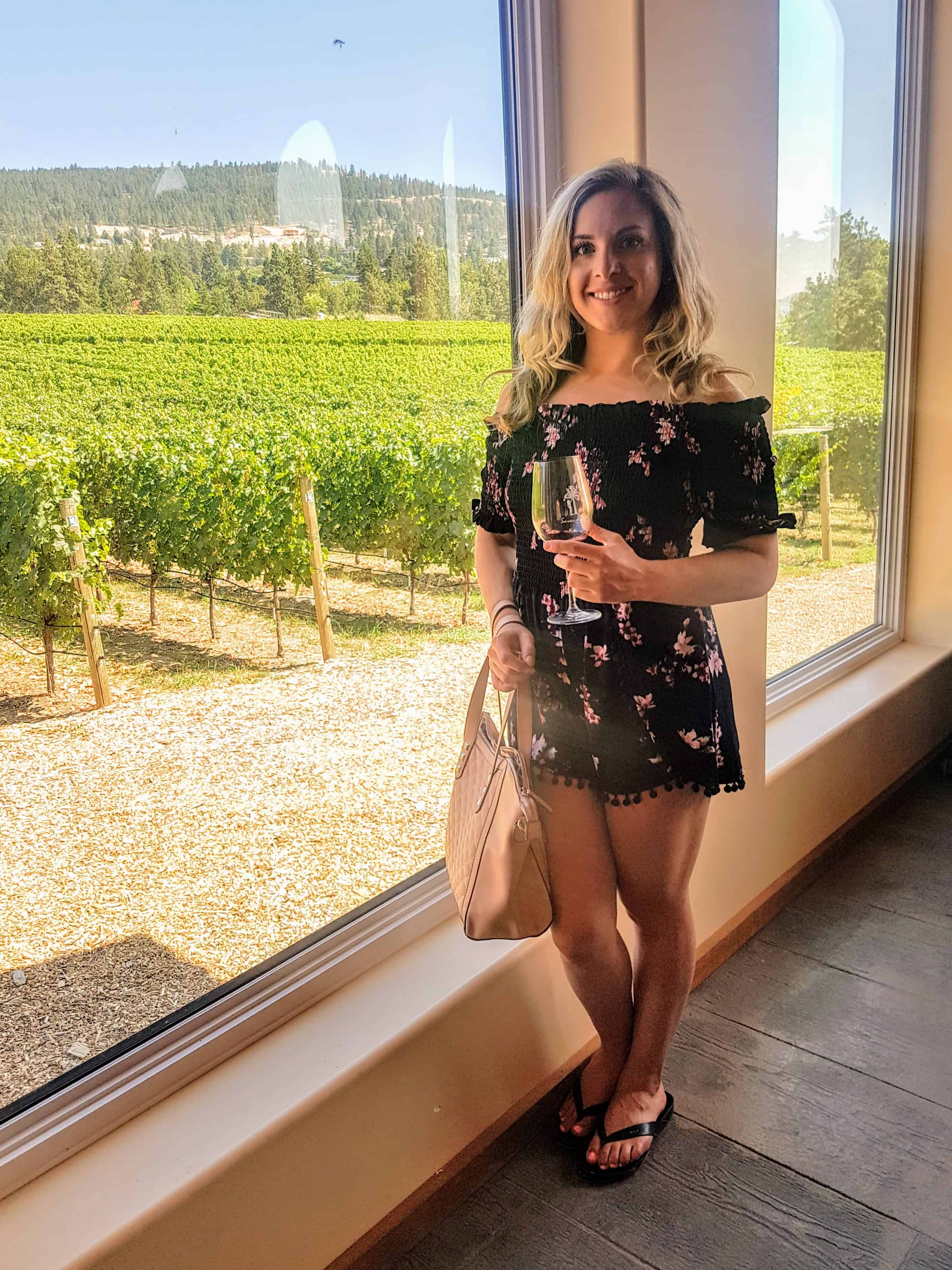 a girl holds a glass of wine standing in front of a window looking out onto the grape vines