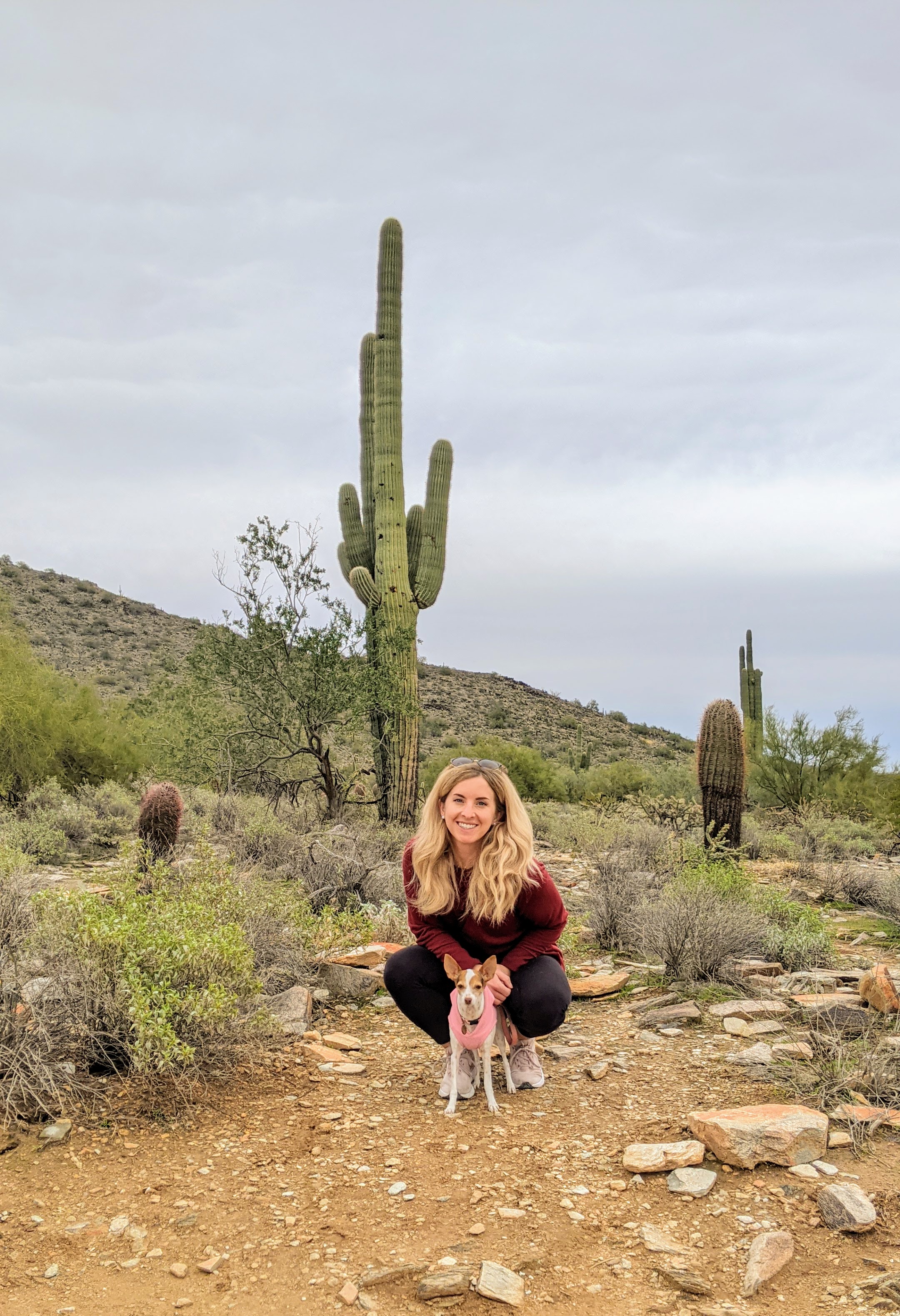 lady poses with her dog on a rocky trail in front of a large cactus